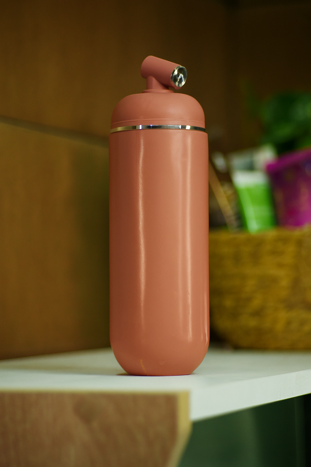 Portable cylindrical bidet bottle in a soft pastel dusty rose shade, sitting on a white shelf along with a basket that contains cosmetic product with a warm indoor background.